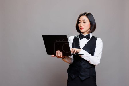 Photo for Hospitality service receptionist in uniform working on laptop and typing on keyboard in studio. Young attractive asian waitress holding portable computer and managing orders online - Royalty Free Image