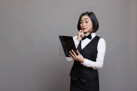 Photo for Thoughtful asian waitress in professional uniform managing order on digital tablet. Pensive receptionist thinking while examining restaurant menu on portable gadget and posing in studio - Royalty Free Image