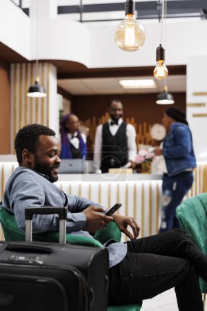 Téléchargez les photos : Afro-américain millenial gars touriste assis avec des bagages dans le hall d'hôtel confortable regardant l'écran du smartphone, en utilisant un téléphone portable à l'échelle internationale, rester connecté avec des amis tout en voyageant, itinérance - en image libre de droit