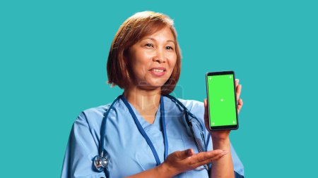 Photo for Professional hospital employee showing instructions video on phone green screen. Asian nurse wearing medical scrubs holding chroma key mock up phone, isolated over studio background, close up - Royalty Free Image