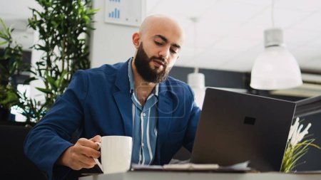 Middle eastern man reads documents online before creating professional marketing report in office. Male worker trying to find business solution, entrepreneur in coworking space.