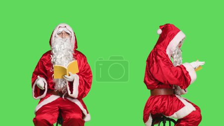 Photo for Saint nick reads lecutre novel on chair, man dressed as santa claus with red suit and glasses while he is reading literature book over greenscreen. Person in costume enjoying fairytale fiction. - Royalty Free Image