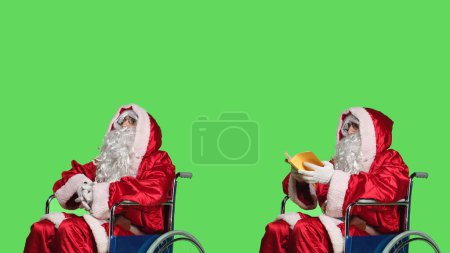 Téléchargez les photos : Père Noël en fauteuil roulant lit livre, dépeignant père Noël avec handicap physique. Adulte en costume festif lisant poésie ou livre de littérature, jouissant d'un nouveau passe-temps pour la culture et l'éducation. - en image libre de droit