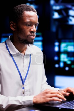 Téléchargez les photos : Développeur informatique afro-américain dans le centre de données à l'aide d'un ordinateur portable pour mettre en place des outils de gestion de configuration qui permettent un basculement automatique et un équilibrage de charge, empêchant le plantage du système des armoires serveur - en image libre de droit