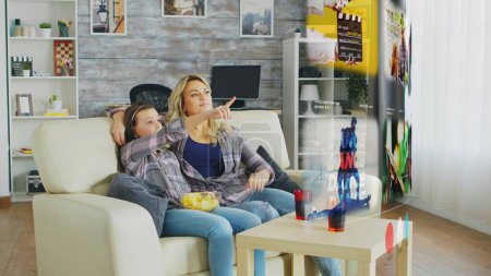 Photo for Parent and child relaxing in living room, watching online content on streaming services while enjoying snacks and beverages. Bored family picking what TV program to watch on media projector - Royalty Free Image