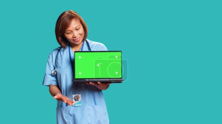 Téléchargez les photos : Infirmière joyeuse montrant des instructions médicales vidéo sur ordinateur portable maquette chroma écran vert clé. Employé de l'hôpital présentant bande de soins de santé explicative sur gadget, isolé sur fond bleu studio - en image libre de droit