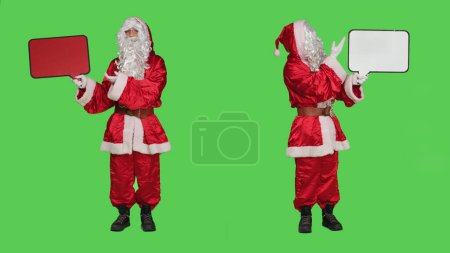 Photo for Saint nick holding speech bubble red billboard while he wears festive traditional costume and hat. Santa embodiment showing cardboard icon for advertisement, full body greenscreen. - Royalty Free Image
