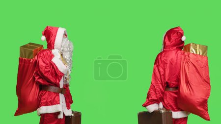 Téléchargez les photos : Nick Saint avec sac et valise portant des cadeaux pour la fête de Noël, fond d'écran vert. Jeune homme dépeignant célèbre personnage d'hiver avec mallette et boîtes-cadeaux. - en image libre de droit