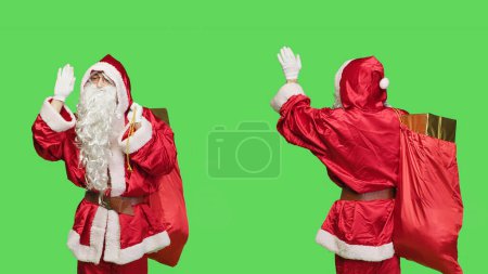 Photo for Smiling santa greeting people on camera, waving hello and acting cheerful feeling happy spreading holiday spirit. Father christmas with bag full of gifts boxes celebrating seasonal winter event. - Royalty Free Image