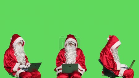 Foto de Moderno Santa Claus funciona en el ordenador portátil, fondo de pantalla verde en el estudio. Padre Navidad sentado en la silla y el uso de PC inalámbrico para la navegación en línea del sitio web, en busca de direcciones para niños. - Imagen libre de derechos
