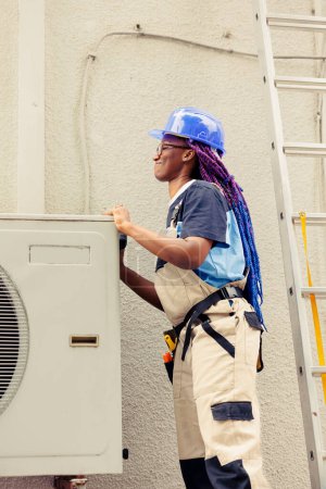 Photo for African american engineer dissasembling outdoor air conditioner metal top coil panel to check for malfunctioning components. Certified technician doing repairing on damaged hvac system - Royalty Free Image