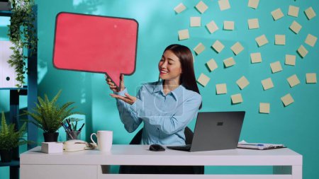 Photo for Happy cheerful asian businesswoman holding red speech bubble sign of empty space for text and showing ok hand gesture. Smiling employee presenting thought bubble cardboard in colourful workplace - Royalty Free Image