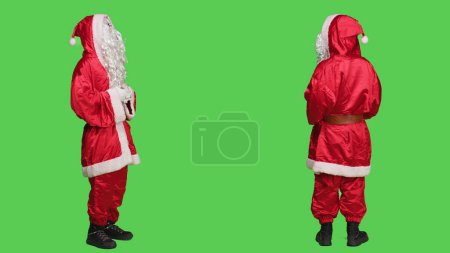 Photo for Saint nick character laughing on camera, saying ho ho ho and spreading christmas spirit during winter holidays. Young man dressed as santa with red costume and hat, full body greenscreen. - Royalty Free Image