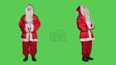 Foto de Saint nick persona en traje rojo festivo riendo sobre pantalla verde de cuerpo completo, haciéndose pasar por personaje principal de temporada durante la celebración del invierno. Adulto retratando a Santa Claus con barba blanca y sombrero. - Imagen libre de derechos