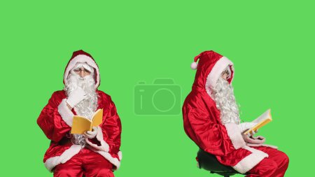 Photo for Father christmas with a reading hobby, enjoying lecture with poetry book or novel story while he sits on chair against greenscreen studio. Santa claus embodiment read literature or fiction. - Royalty Free Image