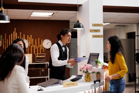 Photo for Hotel guest receiving registration form from helpful asian male concierge. Relaxed asian middle aged woman on vacation making necessary preparation for check in at modern resort - Royalty Free Image