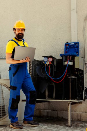 Photo for Portrait of smiling knowleadgeable engineer working on external hvac system, holding laptop. Professional wireman optimizing condenser performance, ensuring it operates at maximum capacity - Royalty Free Image