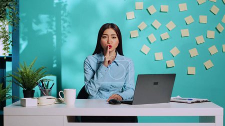 Photo for Stern serious businesswoman shushing at camera, holding quiet gesture finger to lips. Strict pensive office employee at bright colourful modern workplace over blue studio background - Royalty Free Image