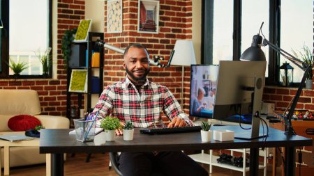 Téléchargez les photos : Heureux souriant homme afro-américain travaillant de la maison, assis au bureau dans un élégant appartement confortable avec un design intérieur éclectique hygge créatif. Joyeux télétravailleur optimiste faisant du travail à distance, gros plan - en image libre de droit