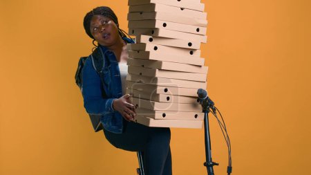 Photo for Youthful black woman holding stack of pizza boxes for delivering to a celebration event in neighborhood. African american delivery person cautiously balancing huge order for food delivery. - Royalty Free Image