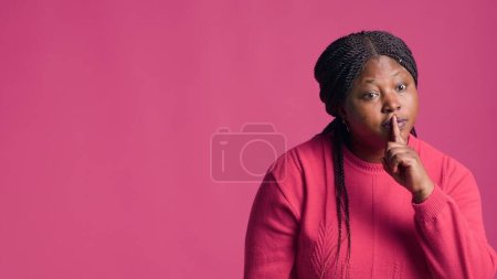 Téléchargez les photos : Femme afro-américaine se tient devant fond rose en utilisant des mouvements de main pour s'exprimer. blogueur femelle dirige l'attention en plaçant doucement le doigt sur les lèvres pour signaler la tranquillité. - en image libre de droit