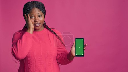 Photo for Female african american beauty vertically holds mobile device displaying green screen. Stunning black woman showing innovative online shopping app on smartphone with blank mockup copyspace template. - Royalty Free Image
