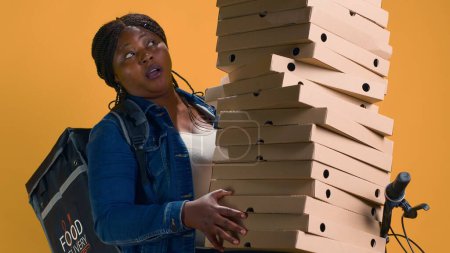 Téléchargez les photos : Livreur afro-américain équilibrant beaucoup de boîtes à pizza pour livrer au quartier. Femme noire active à vélo portant soigneusement une grosse commande de nourriture pour le transport. - en image libre de droit