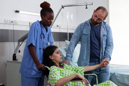 Foto de Enfermera ginecóloga ayudando a la mujer embarazada a ir a cirugía en la sala de hospital. Paciente con embarazo sentada en silla de ruedas siendo reconfortante por marido y asistente en maternidad - Imagen libre de derechos