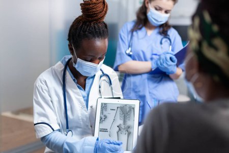 Photo for Bone doctor explaining clinical information on tablet to patient during orthopedic medical exam. Orthopedist in modern sterile hospital office wearing face mask and medical gloves - Royalty Free Image