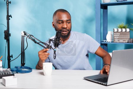 Photo for Internet influencer speaking in microphone and using laptop while recording video for blog in home studio. African american vlogger streaming live talking and reading from computer in mic - Royalty Free Image
