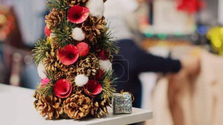 Photo for Extreme close up shot of festive miniature Christmas tree adorned with pine cones and flowers sitting on clothing store counter during winter holiday season. Xmas decor in fashion boutique - Royalty Free Image