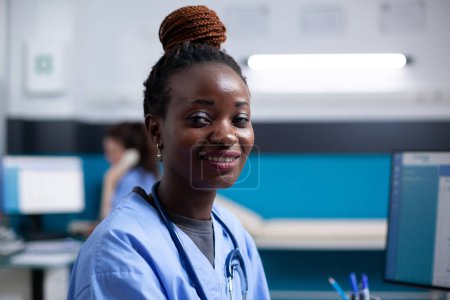 Téléchargez les photos : Infirmière afro-américaine souriante à l'intérieur d'un bureau médical moderne et propre. Heureuse femme adulte professionnelle portant des blouses uniforme et stéthoscope, regardant la caméra, portrait en gros plan - en image libre de droit