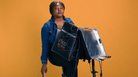 Photo for Efficient delivery woman unloading fast food takeway after arrival with bicycle in customer neighborhood. African american courier unpacking her food delivery bag at client destination. - Royalty Free Image