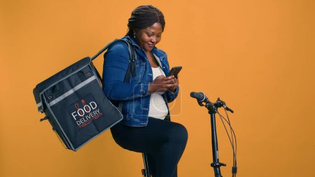 Photo for African american courier carrying packages from local restaurants on bike as eco-friendly transportation. Woman with smartphone ensures efficient deliveries while reducing environmental impact. - Royalty Free Image
