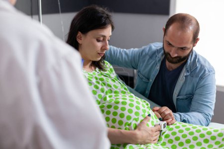 Foto de Ginecólogo revisando los síntomas de la mujer embarazada antes de la cirugía médica, discutiendo el proceso de parto con los futuros padres en la sala de hospital. Paciente con embarazo acostada en cama en clínica de maternidad - Imagen libre de derechos