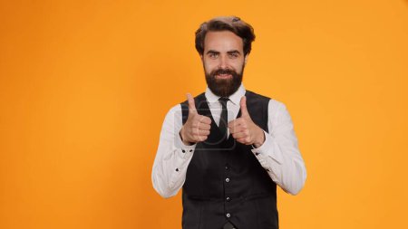 Photo for Happy server gives like symbol on camera, expressing his approval with okay gesture. Professional restaurant employee giving thumbs up to show positive sign, working in catering industry. - Royalty Free Image
