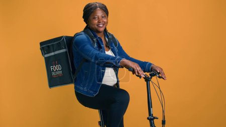 Téléchargez les photos : Femme afro-américaine active à vélo avec sac sur le dos va livrer de la nourriture. Jeune femme noire portant un sac de livraison de nourriture prend des commandes aux clients. - en image libre de droit