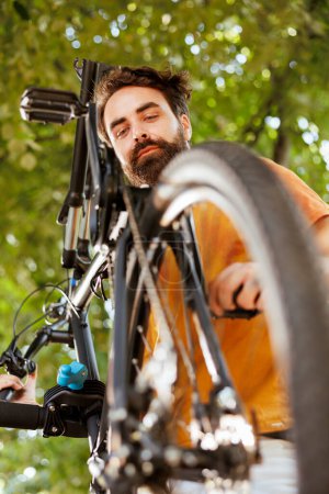 Photo for Healthy vibrant caucasian man is seen outdoors doing annual upkeeping and maintenance of modern bicycle. Active young male cyclist checking and ensuring functioning of bike pedal. - Royalty Free Image