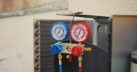 Photo for Dolly in close up of yellow safety helmet and manifold gauges used for checking air conditioner freon in need of maintenance. Set of refrigerant levels benchmarking tool mounted on condenser - Royalty Free Image
