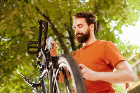 Photo for Active and healthy male cyclist inspecting his bikes wheel for damages in the yard. Image portrays young caucasian man using lubricant to fix broken component ensuring a secure and safe ride - Royalty Free Image