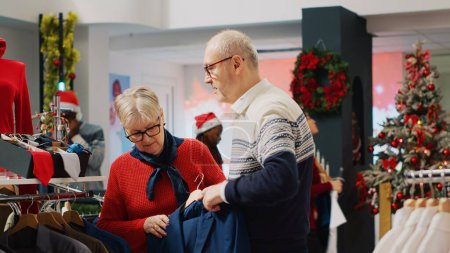 Photo for Woman browsing through xmas adorn clothing store racks with husband in Christmas shopping spree. Elderly couple searching for elegant attire garments as gift for son during winter holiday season - Royalty Free Image