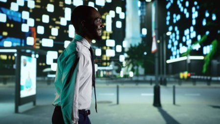 Foto de Hombre afroamericano camina por el centro por la noche y admira los rascacielos urbanos modernos bajo las luces de la ciudad. Persona alegre en traje de moda caminando cerca del distrito de negocios en el centro de la ciudad. - Imagen libre de derechos
