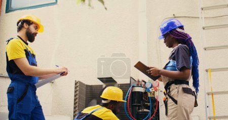 Photo for Engineers cleaning outdoor condenser internal coolants on a sunny summer day, checking freon level and calibrating thermostats while colleague checks maintenance procedures on tablet - Royalty Free Image