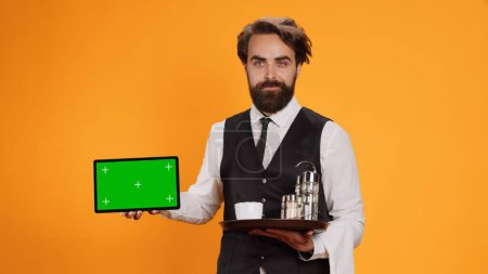Photo for Smiling butler presents greenscreen on tablet, posing with confidence against yellow background. Young adult worker showing isolated template on smartphone display in front of camera. - Royalty Free Image