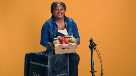 Téléchargez les photos : Coursier afro-américain portant soigneusement panier de fruits du sac de livraison de nourriture. Sourire femme noire livrant des fruits et légumes frais avec vélo comme moyen de transport. - en image libre de droit