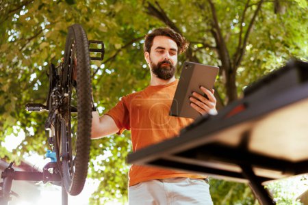 Photo for Active sporty man in his yard using smart digital device to research bicycle repair solutions. Dedicated caucasian male holding his phone tablet and bike checking for damages outdoor. - Royalty Free Image