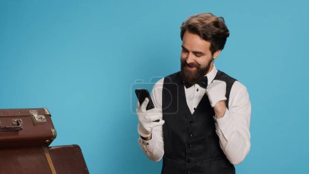 Cheerful bellboy on smartphone app, feeling excited about good news on social media online in studio. Remarkable doorman with white gloves acting positive and joyful against blue background.