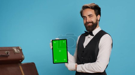 Photo for Stylish bellboy presents tablet in studio and showing blank greenscreen display while he stands next to trolley bags. Classy porter showcasing concierge assistance with luxury service. - Royalty Free Image