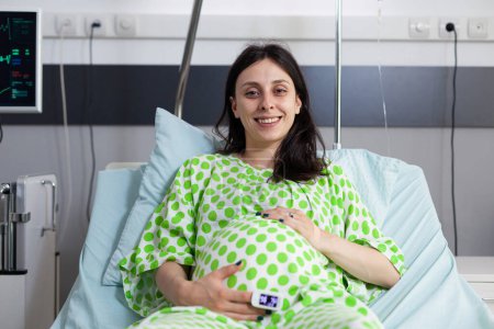 Téléchargez les photos : Patiente enceinte joyeuse attendant que le médecin commence l'examen de santé lors de la visite de contrôle dans la salle d'hôpital. Femme enceinte du ventre couchée au lit, souriant à la caméra avant la chirurgie - en image libre de droit