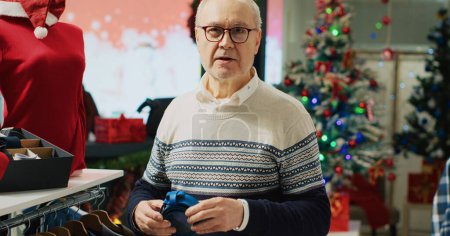 Foto de Cliente anciano mirando cinturones, admirando bellas artesanías mientras va de compras en la tienda decorada de Navidad. Antiguo cliente en el centro comercial boutique de moda en busca de regalos de Navidad para ofrecer a la familia - Imagen libre de derechos